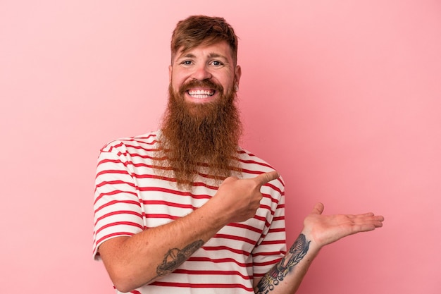 Young caucasian ginger man with long beard isolated on pink background excited holding a copy space on palm.