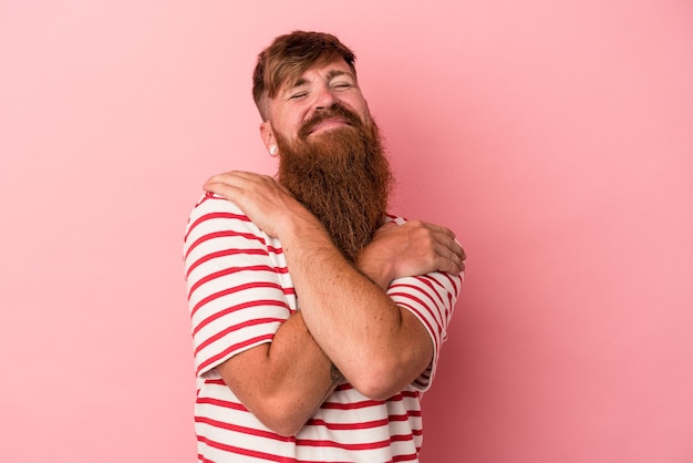 Il giovane uomo caucasico dello zenzero con la barba lunga isolato sugli abbracci rosa del fondo, sorride spensierato e felice.