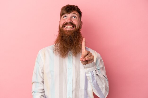 young caucasian ginger man with long beard isolated on pink background indicates with both fore fingers up showing a blank space.