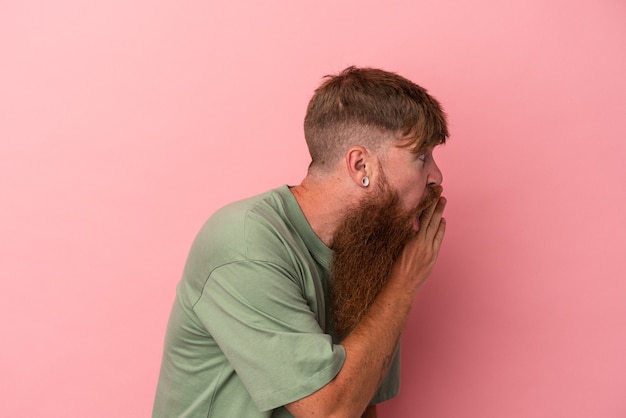 Young caucasian ginger man with long beard isolated on pink background is saying a secret hot braking news and looking aside