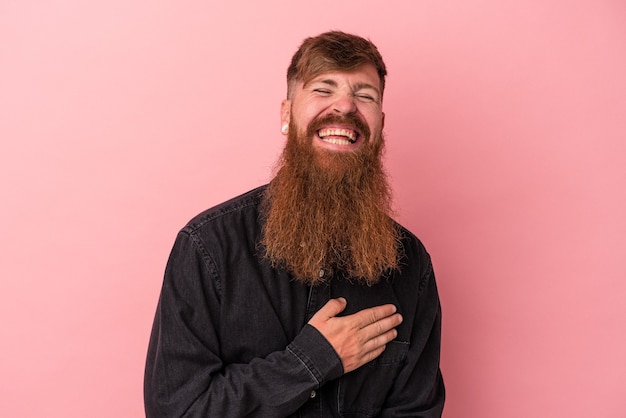 Young caucasian ginger man with long beard isolated on pink background laughing keeping hands on heart, concept of happiness.