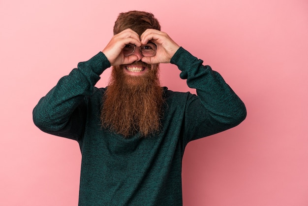 Young caucasian ginger man with long beard isolated on pink background showing okay sign over eyes