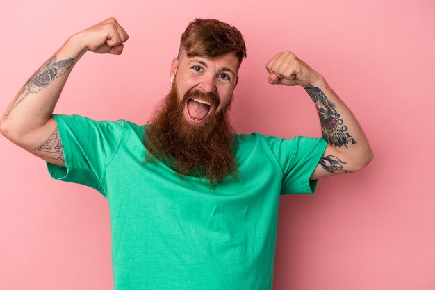 Young caucasian ginger man with long beard isolated on pink background showing strength gesture with arms, symbol of feminine power