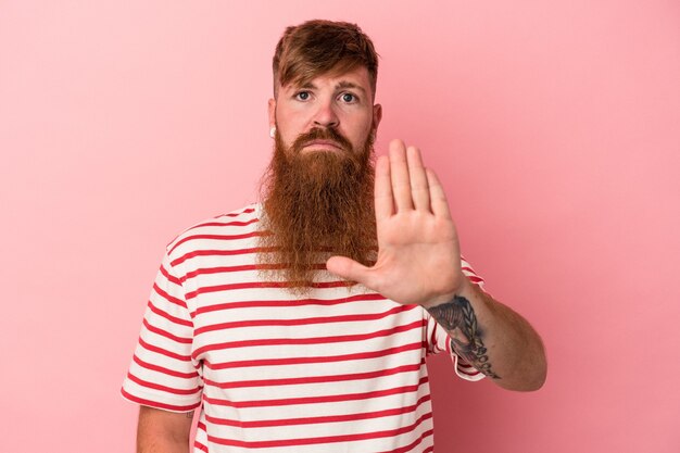 Young caucasian ginger man with long beard isolated on pink background standing with outstretched hand showing stop sign, preventing you.