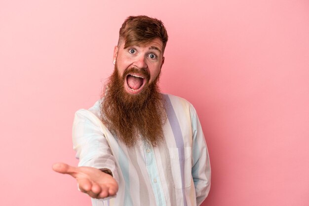 Young caucasian ginger man with long beard isolated on pink background stretching hand at camera in greeting gesture.