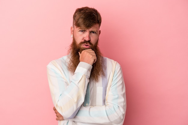 Young caucasian ginger man with long beard isolated on pink background suspicious, uncertain, examining you.