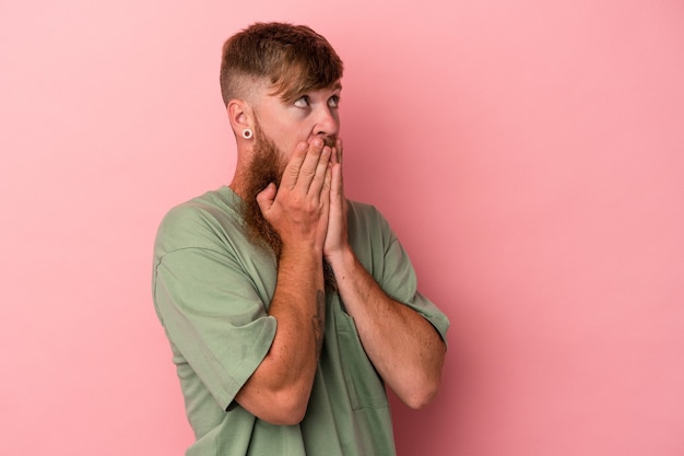 Young caucasian ginger man with long beard isolated on pink background thoughtful looking to a copy space covering mouth with hand