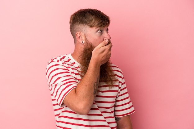 Giovane uomo caucasico dello zenzero con la barba lunga isolato su fondo rosa premuroso che guarda ad uno spazio della copia che copre la bocca con la mano.