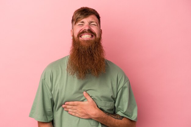 Young caucasian ginger man with long beard isolated on pink background touches tummy, smiles gently, eating and satisfaction concept.