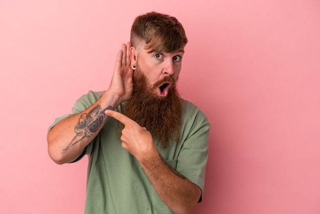 Young caucasian ginger man with long beard isolated on pink background trying to listening a gossip.