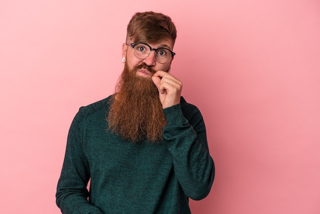 Giovane uomo caucasico dello zenzero con la barba lunga isolata su fondo rosa con le dita sulle labbra che tengono un segreto.