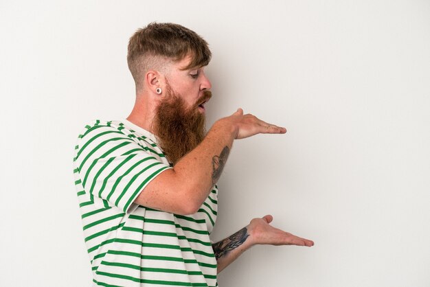 Il giovane uomo caucasico dello zenzero con la barba lunga isolato su fondo bianco ha colpito e stupito che tiene uno spazio della copia fra le mani.