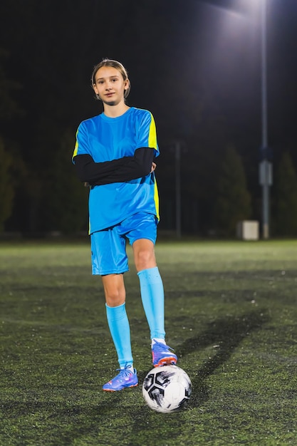 Молодая кавказская девушка в синей спортивной одежде позирует с футбольным мячом на стадионе с полным вертикальным выстрелом