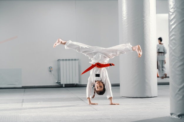 Young Caucasian girl in dobok doing hand stand with spread legs on taekwondo class.