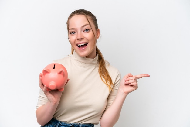 Young caucasian girl holding a piggybank isolated on white background surprised and pointing finger to the side