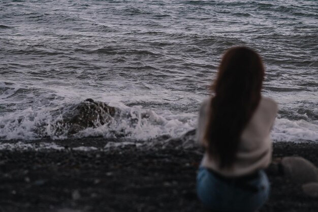 Foto una giovane ragazza caucasica è seduta su una roccia vicino al mare e guardando l'orizzonte un'immagine sfocata tempo inclemente freddo grigio