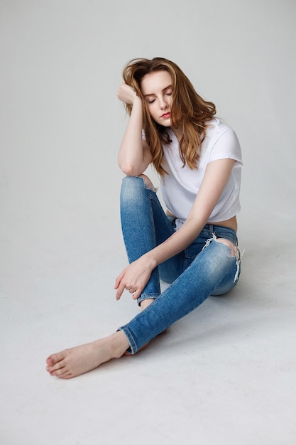 Young caucasian girl posing in t-shirt, ripped jeans, sitting on studio floor