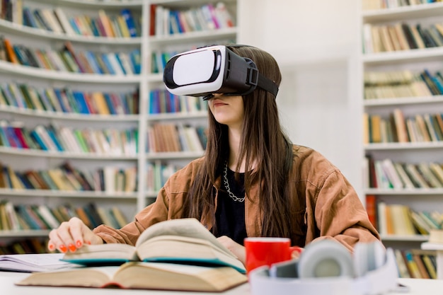 Young Caucasian girl student in brown shirt sitting at the table with books in college library and studying using VR goggles