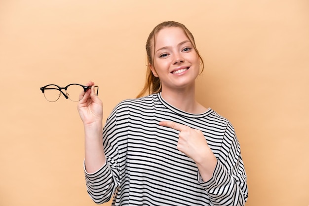 Young caucasian girl with glasses isolated on beige background and pointing it