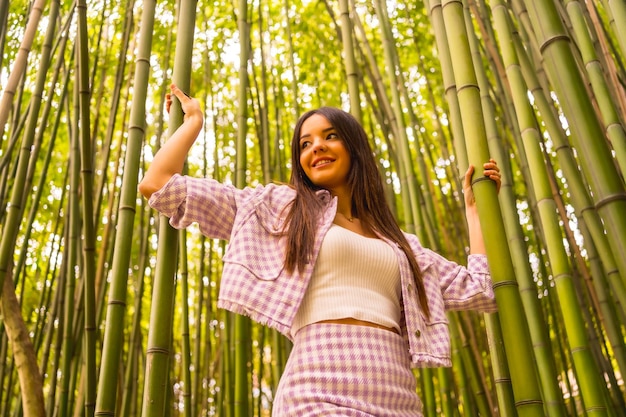 竹林にピンクのスカートをはいた白人少女熱帯気候の夏休みを楽しんで森の中を歩き、竹の幹を掴んで笑う