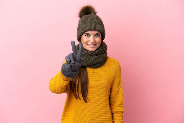 Young caucasian girl with winter hat isolated on pink background smiling and showing victory sign