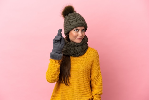 Young caucasian girl with winter hat isolated on pink background with fingers crossing and wishing the best