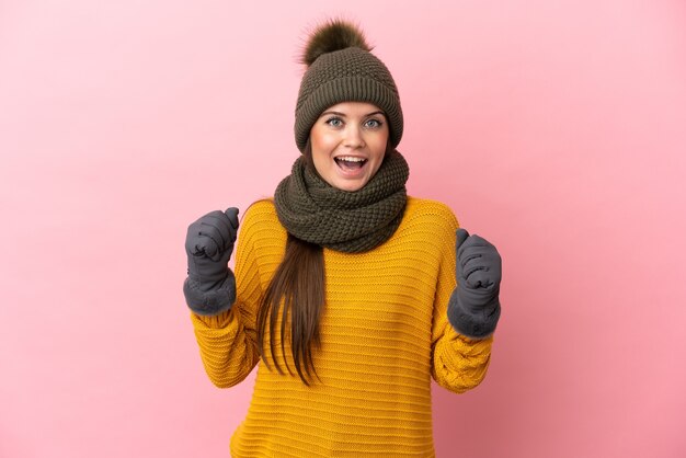 Young caucasian girl with winter hat isolated on pink wall celebrating a victory in winner position