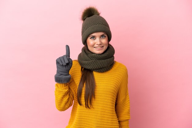 Молодая кавказская девушка в зимней шапке изолирована на розовой стене, показывая и поднимая палец в знак лучших