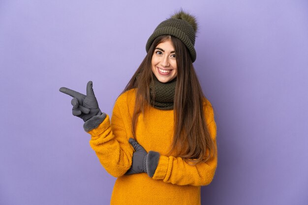 Giovane ragazza caucasica con cappello invernale isolato sulla parete viola che punta il dito a lato