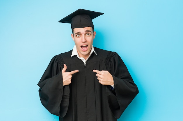 Young caucasian graduated man surprised pointing at himself, smiling broadly.