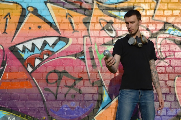 Young caucasian graffiti artist in black t-shirt with silver aerosol spray can near colorful graffiti in pink tones on brick wall. Street art and contemporary painting process