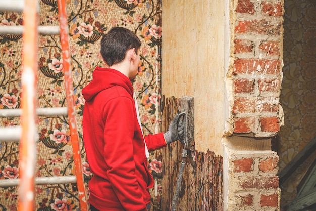 Photo a young caucasian guy removes retro wallpaper using a steamer machine in an old abandoned house