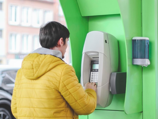 A young caucasian guy in a yellow jacket stands near a green ATM at a gas station and pays for gasoline