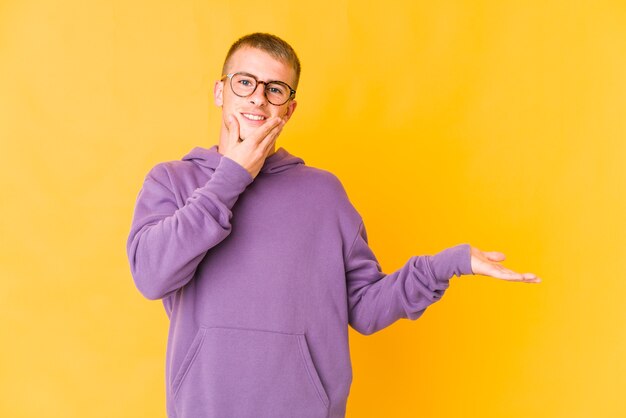 Young caucasian handsome man holds copy space on a palm, keep hand over cheek. Amazed and delighted.