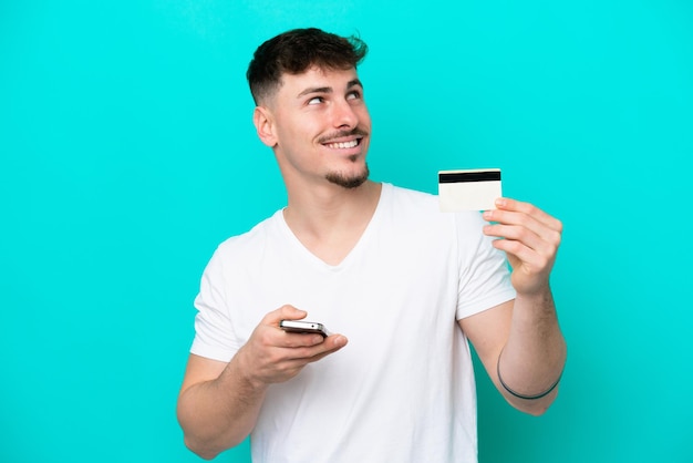 Young caucasian handsome man isolated on blue background buying with the mobile with a credit card while thinking