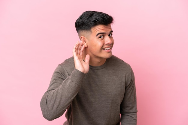 Young caucasian handsome man isolated on pink background listening to something by putting hand on the ear