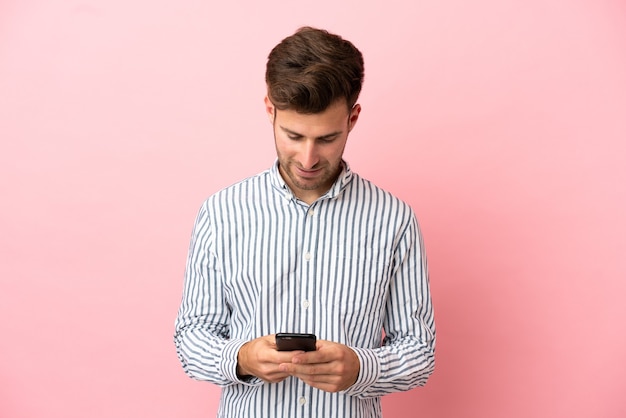 携帯電話でメッセージを送信するピンクの背景に分離された若い白人ハンサムな男