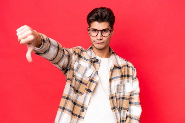 Young caucasian handsome man isolated on red background showing thumb down with negative expression