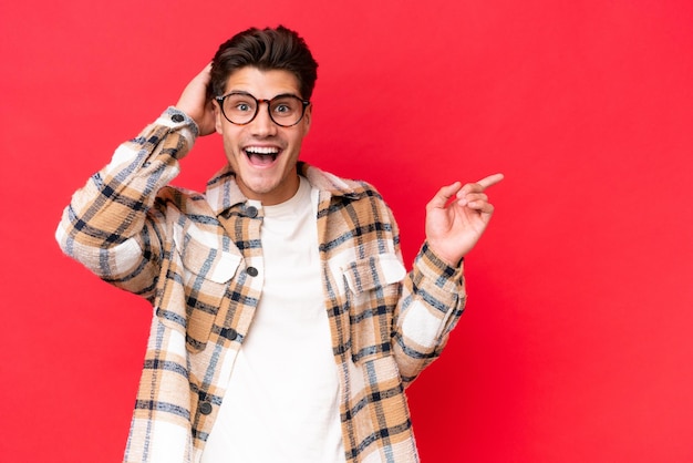 Young caucasian handsome man isolated on red background surprised and pointing finger to the side