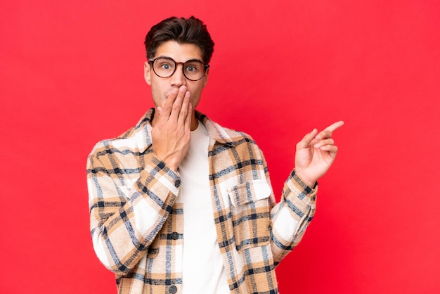 Young caucasian handsome man isolated on red background with surprise expression while pointing side