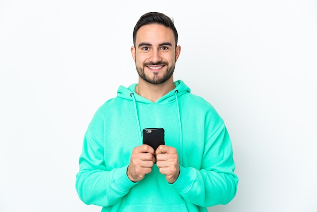 Giovane uomo caucasico bello isolato su sfondo bianco guardando la telecamera e sorridente mentre si utilizza il cellulare