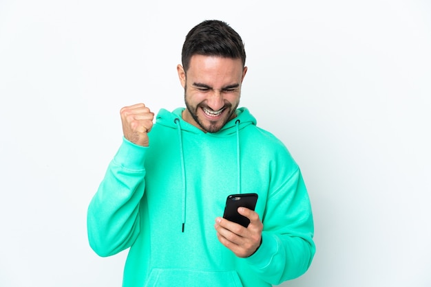 Young caucasian handsome man isolated on white wall using mobile phone and doing victory gesture