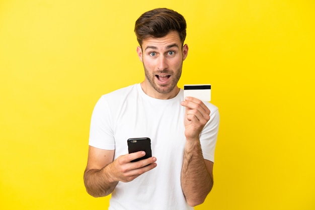 Young caucasian handsome man isolated on yellow background buying with the mobile and holding a credit card with surprised expression