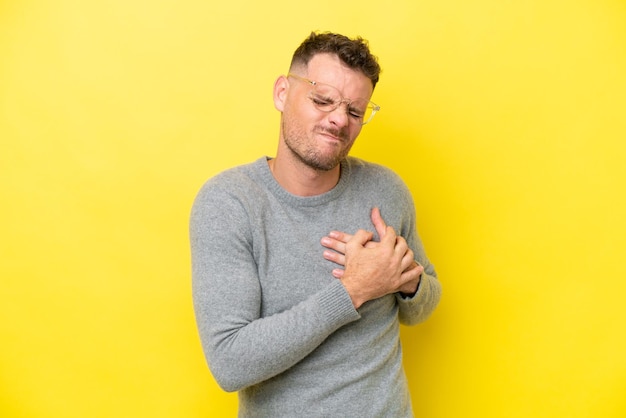 young caucasian handsome man isolated on yellow background having a pain in the heart