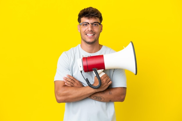 Young caucasian handsome man isolated on yellow background holding a megaphone and smiling