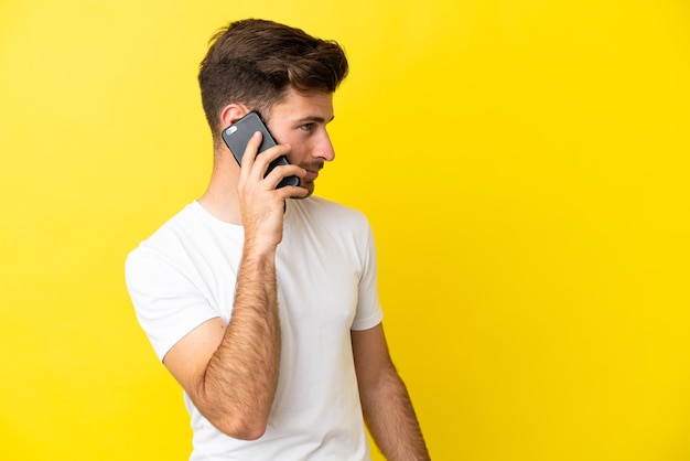 Young caucasian handsome man isolated on yellow background keeping a conversation with the mobile phone with someone