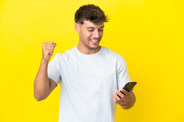 Young caucasian handsome man isolated on yellow background using mobile phone and doing victory gesture