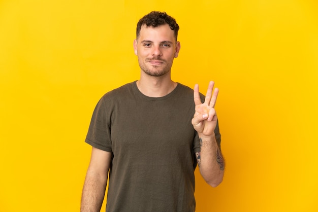 Young caucasian handsome man isolated on yellow wall happy and counting three with fingers