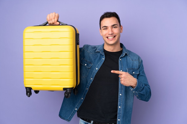 Young caucasian handsome man on purple wall in vacation with travel suitcase