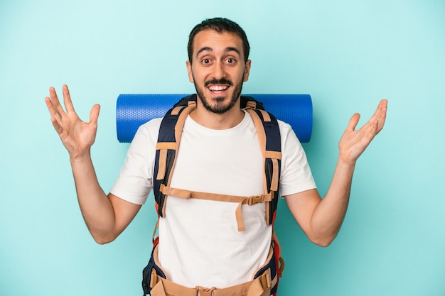 Young caucasian hiker man isolated on blue background receiving a pleasant surprise, excited and raising hands.